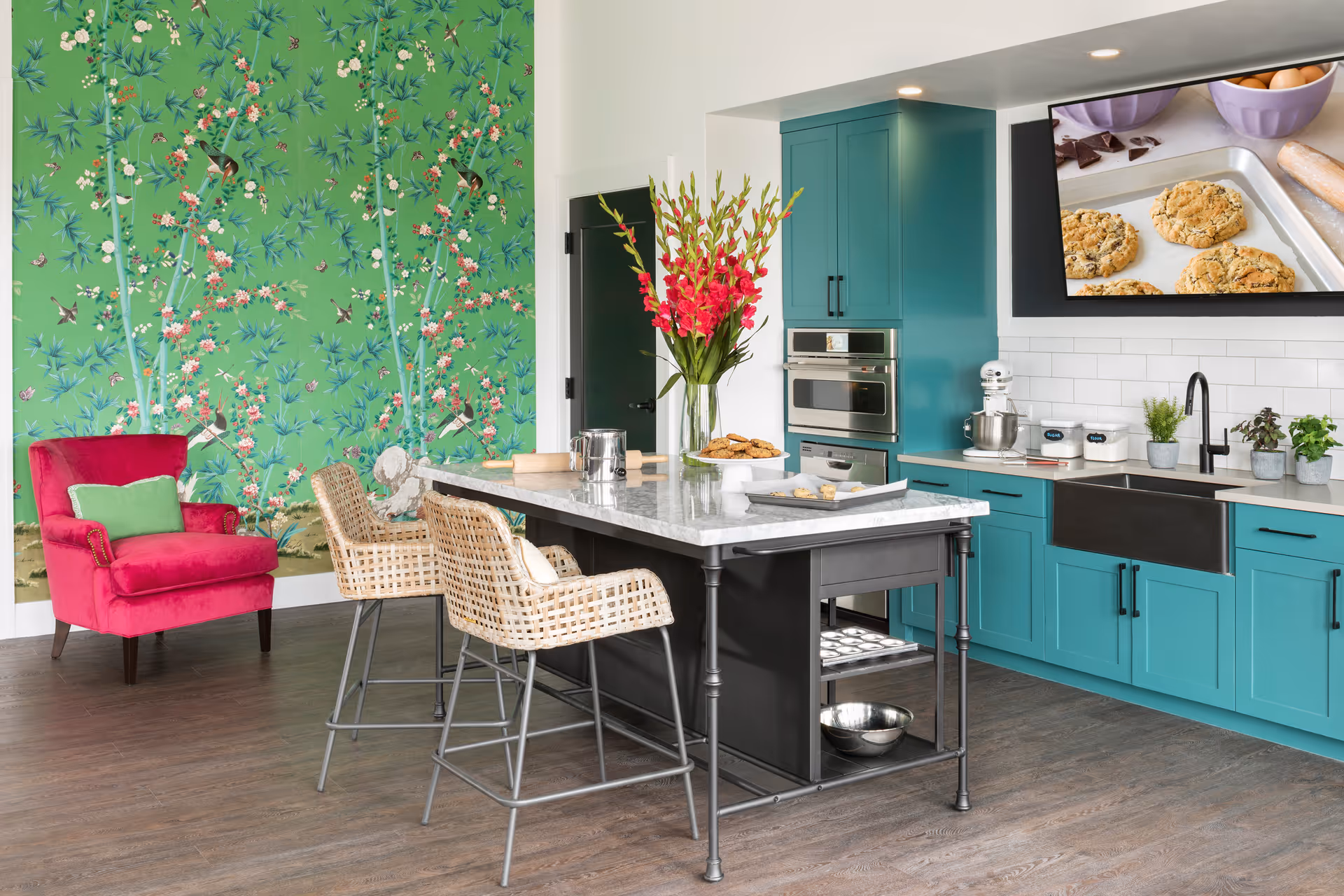 Bright communal kitchen with teal cabinets, a marble-topped island and wicker bar stools beside a green floral accent wall and a pink armchair.