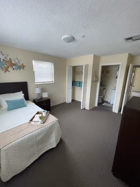 A clean and bright bedroom with a bed featuring white bedding and a beige throw, a nightstand with a lamp and decorative items, a window with blinds, an open closet with shelves and hanging space, and a doorway leading to a bathroom.