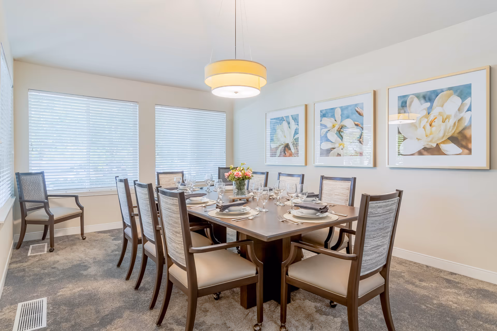A bright dining room with a rectangular wooden table set for eight people. The table is arranged with plates, silverware, glasses, and napkins, and a vase with flowers is placed in the center. The room has three large windows with white blinds, three framed floral artworks on the wall, and a modern round ceiling light fixture above the table. The floor is carpeted in a neutral tone.