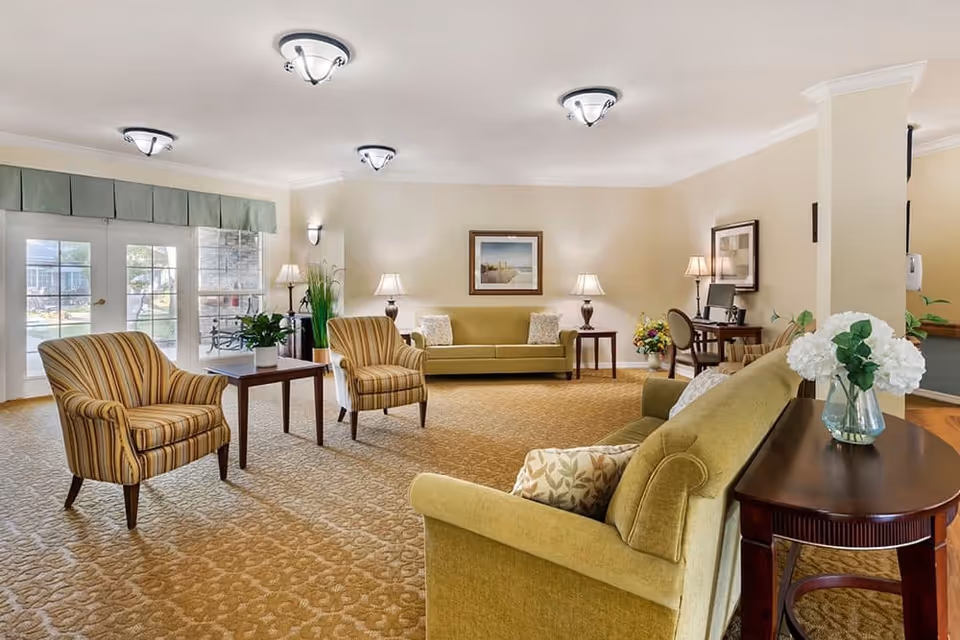 A spacious and well-lit living room area with beige walls and carpet. The room features two striped armchairs, two green sofas, a wooden side table with a vase of white flowers, several table lamps, framed artwork on the walls, and a desk with a computer in the corner. Large windows and glass doors allow natural light to enter, and there are plants placed around the room.