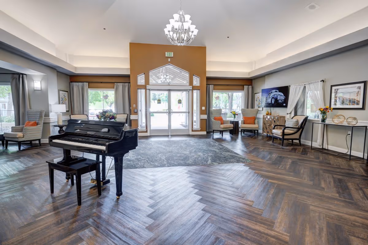 A spacious and well-lit senior living facility common area featuring a black grand piano with a matching bench in the center. The room has large windows with curtains, several comfortable armchairs with orange cushions, a TV mounted on the wall, decorative flowers, and a chandelier hanging from the ceiling. The floor has a herringbone wood pattern with a large area rug near the entrance doors.