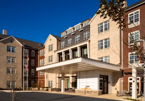 Exterior view of Tribute at Black Hill senior living facility showing a multi-story building with beige and brown brick facade, large windows, and a covered entrance area under a clear blue sky.
