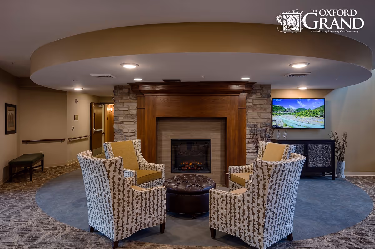 A cozy seating area in a senior living facility featuring four patterned armchairs arranged around a round, tufted ottoman in front of a wooden fireplace with stone accents. A flat-screen TV is mounted on the wall to the right, displaying a scenic landscape. The room has soft lighting and neutral-colored walls and carpet.
