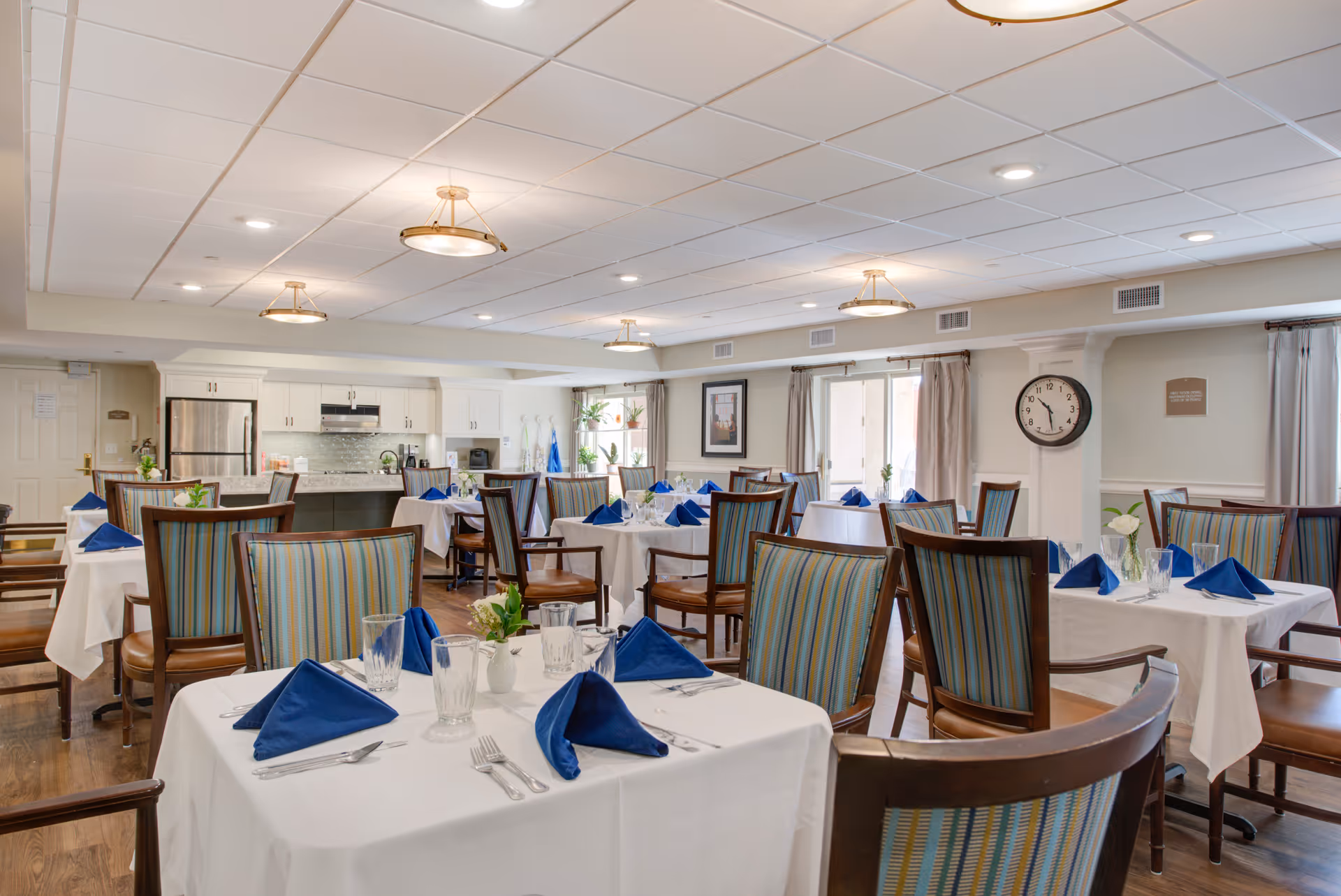 A bright and clean dining room with multiple tables covered in white tablecloths, each set with blue folded napkins, glassware, and silverware. The room features wooden chairs with striped upholstery, a kitchen area with stainless steel appliances in the background, and large windows with curtains allowing natural light to enter.