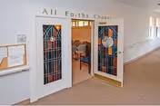Entrance to the All Faiths Chapel with double doors featuring stained glass panels. Inside, rows of chairs are visible. The hallway outside the chapel has carpeted flooring and windows along the right side.