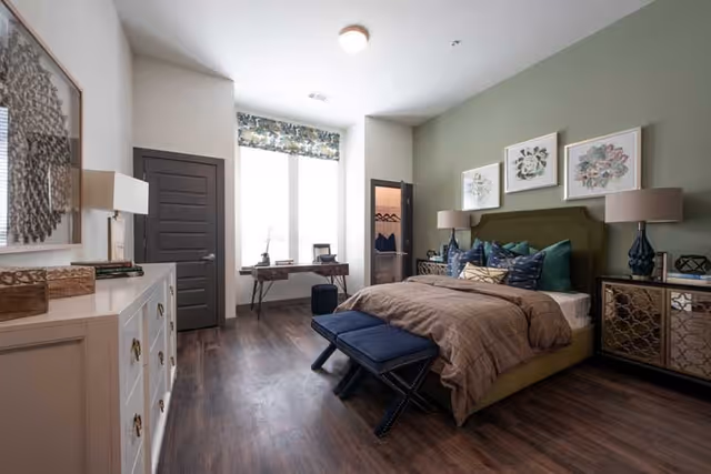 A well-decorated bedroom with a large bed featuring multiple pillows and a brown comforter. There are two blue benches at the foot of the bed, nightstands with lamps on either side, and framed artwork on the green accent wall behind the bed. A dresser with decorative items is on the left side, and a window with a patterned valance lets in natural light. A small desk with a chair is positioned in front of the window, and a door to a closet is visible.