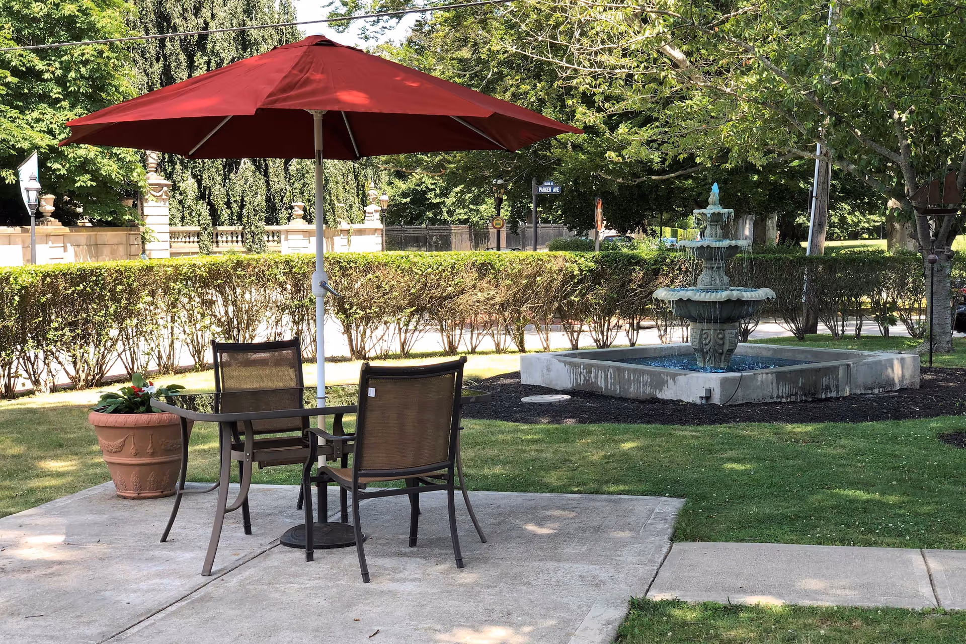 Outdoor patio area with a glass table and two chairs under a large red umbrella. Nearby is a decorative water fountain surrounded by grass and trees, with a hedge and street signs visible in the background.