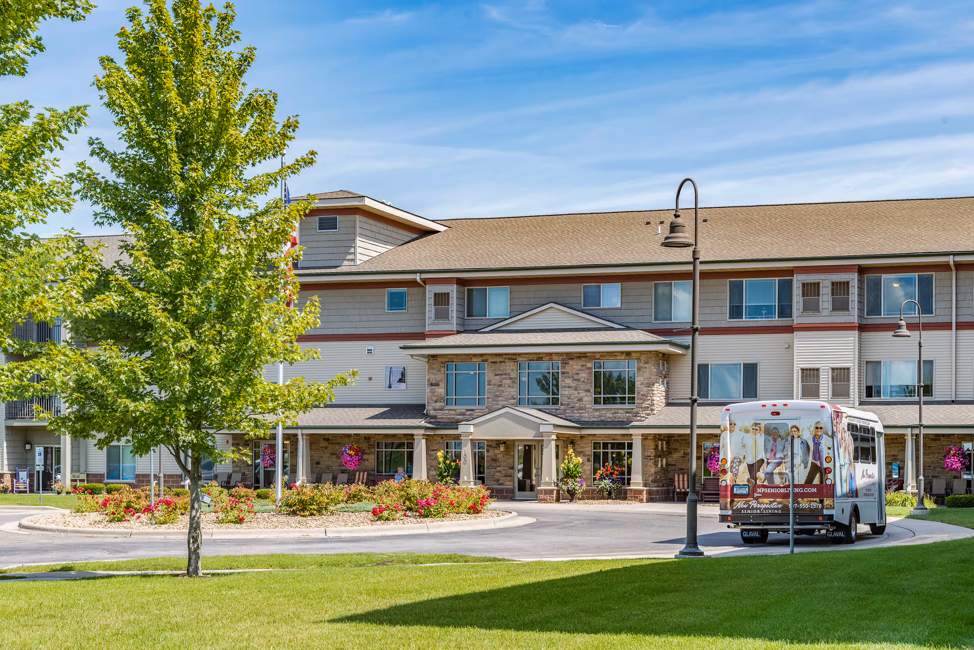 Exterior view of New Perspective Senior Living facility in Mankato, showing a three-story building with a covered entrance, landscaped garden with flowers and trees, a circular driveway, and a shuttle bus parked near the entrance.