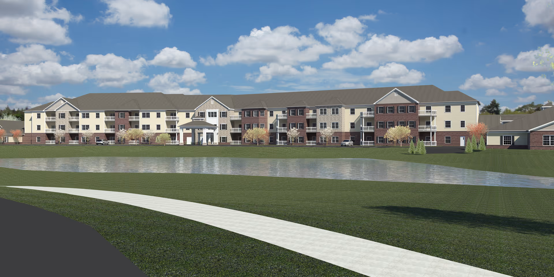 A large, multi-story senior living facility building named Springwood Hudson, viewed from across a pond with a curved walking path in the foreground. The building has a mix of beige and brick exterior walls, multiple windows, and a central entrance with a covered portico. The sky is blue with scattered clouds.