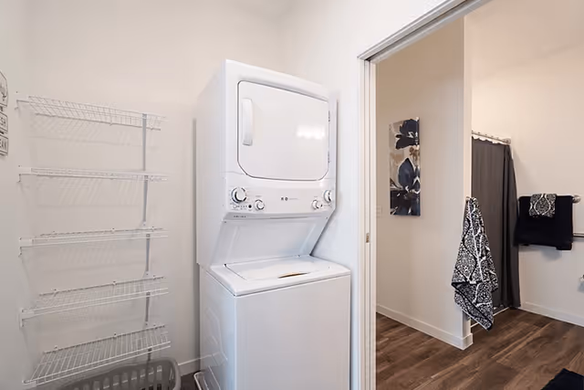 Laundry area with a stacked white washer and dryer unit next to a wall-mounted wire rack and a laundry basket on the floor. Adjacent room visible with a bathroom featuring a shower curtain, towel rack with towels, and a floral wall decoration.