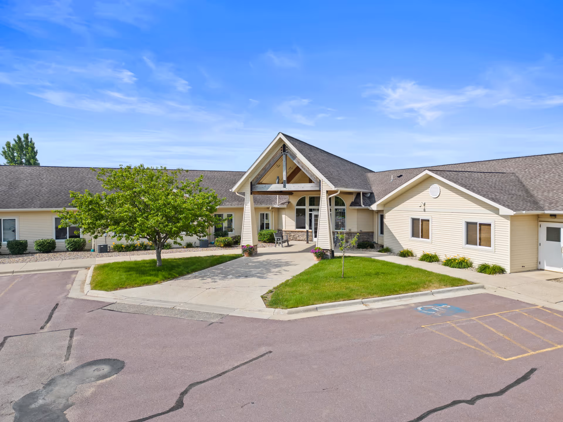 Single-story beige senior living building with a covered entrance, small grassy island and adjacent parking lot under a blue sky.