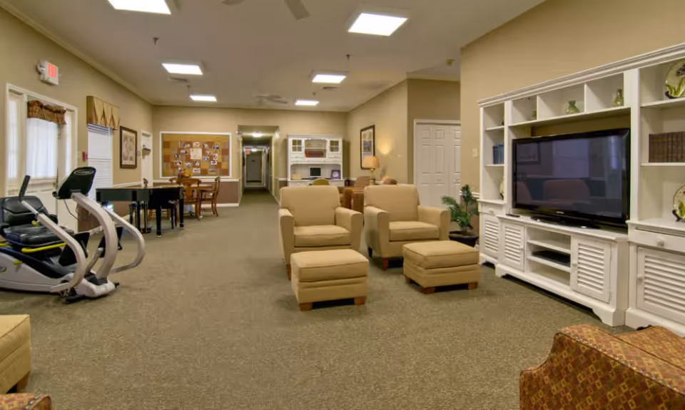 A spacious senior living common area with beige armchairs and matching ottomans facing a large flat-screen TV on a white entertainment center. To the left, there is exercise equipment and a piano near a window with curtains. The room has carpeted floors, neutral-colored walls, and ceiling lights. In the background, there is a hallway and a dining area with a table and chairs.