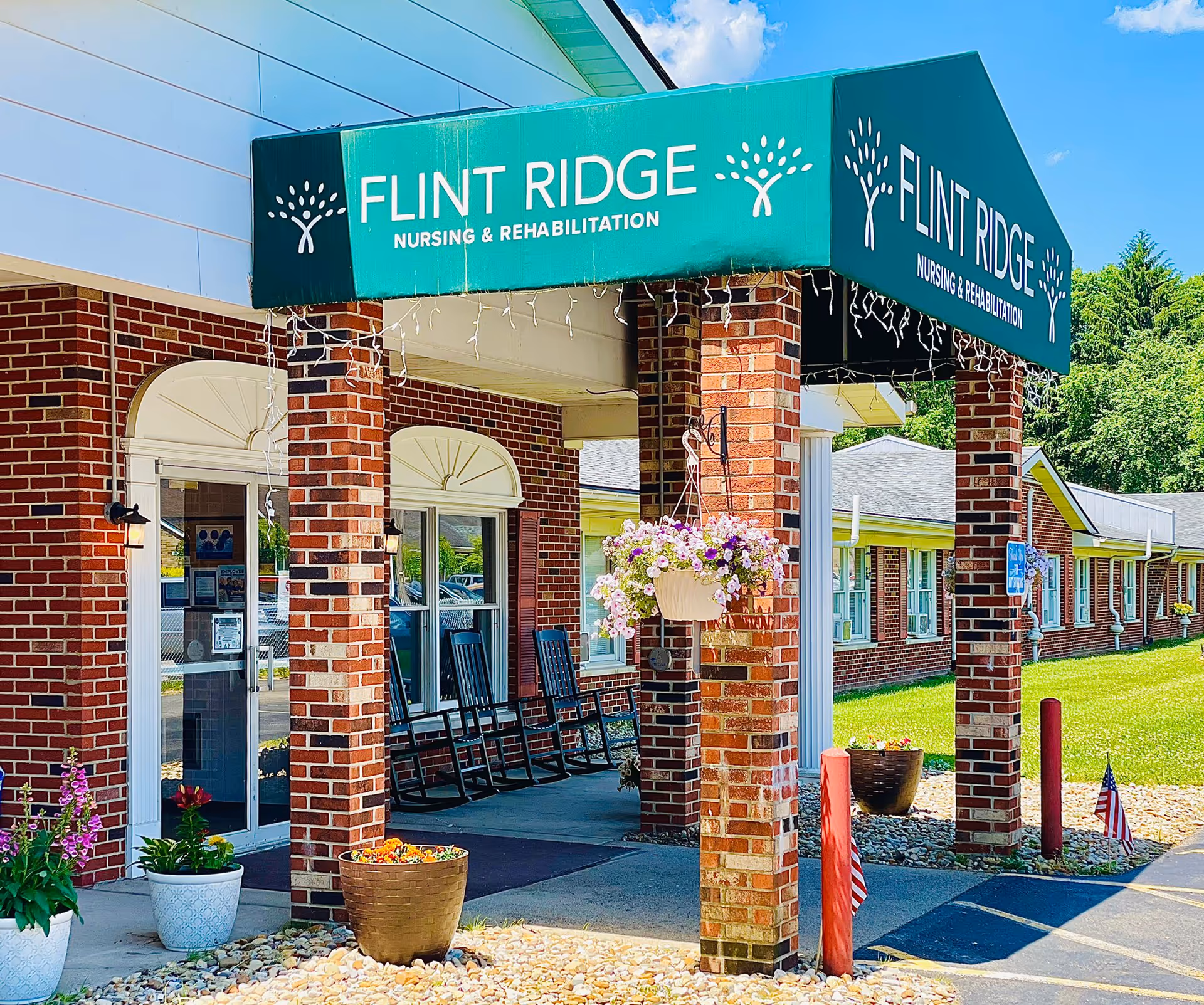Entrance of Flint Ridge Nursing and Rehabilitation facility with a green awning displaying the facility name. The building is made of red brick with white trim, and there are rocking chairs on the covered porch. Flower pots and hanging flower baskets decorate the entrance area under a clear blue sky.