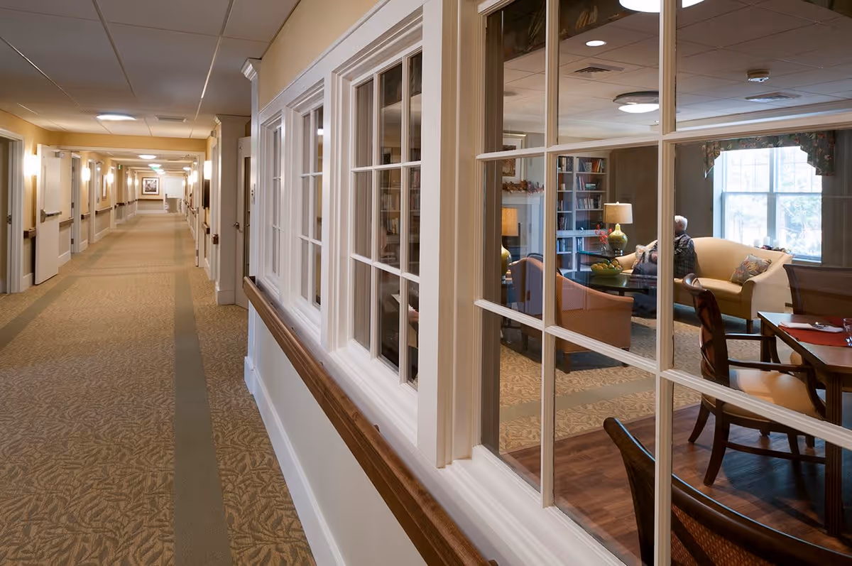 A long, well-lit hallway with beige walls and carpeted floor in a senior living facility. On the right side, large windows reveal a cozy sitting area with armchairs, a sofa, a table with a lamp, bookshelves, and a person seated near a window.