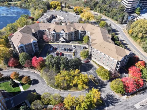 Aerial view of a large senior living facility named Valley View Cooperative surrounded by trees with autumn foliage. The building is U-shaped with multiple floors and a parking lot in the center. There is a pond visible in the upper left corner and roads encircle the property.
