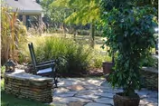 Outdoor garden area with a stone-paved patio, a stone bench with a black chair, surrounded by green plants and trees under sunlight.