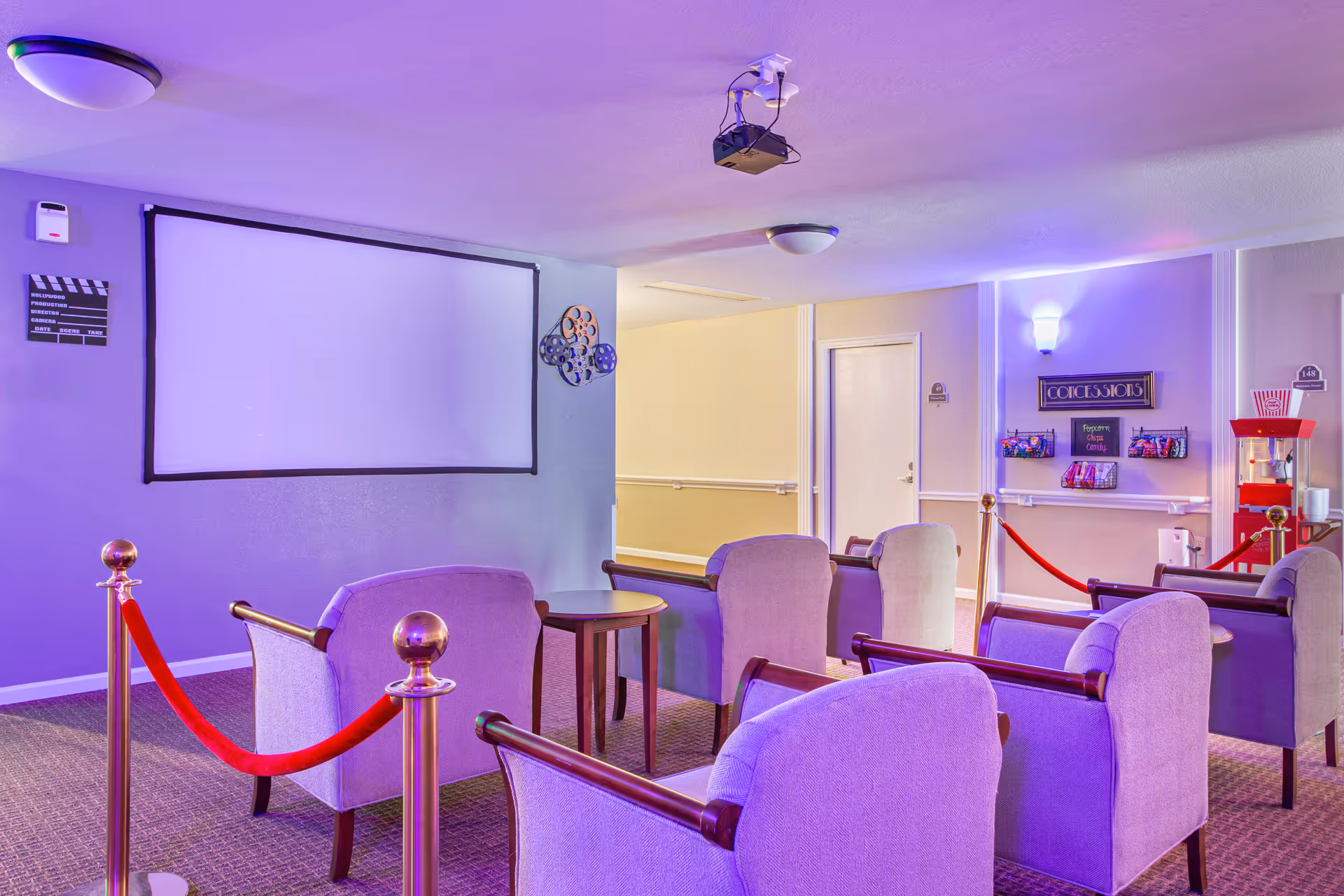 A small movie theater room with several upholstered armchairs arranged in rows facing a large blank projection screen. The room has a popcorn machine and a concession stand with snacks on the right side. The walls are decorated with film reels and a clapperboard. Purple lighting gives the room a cozy ambiance.