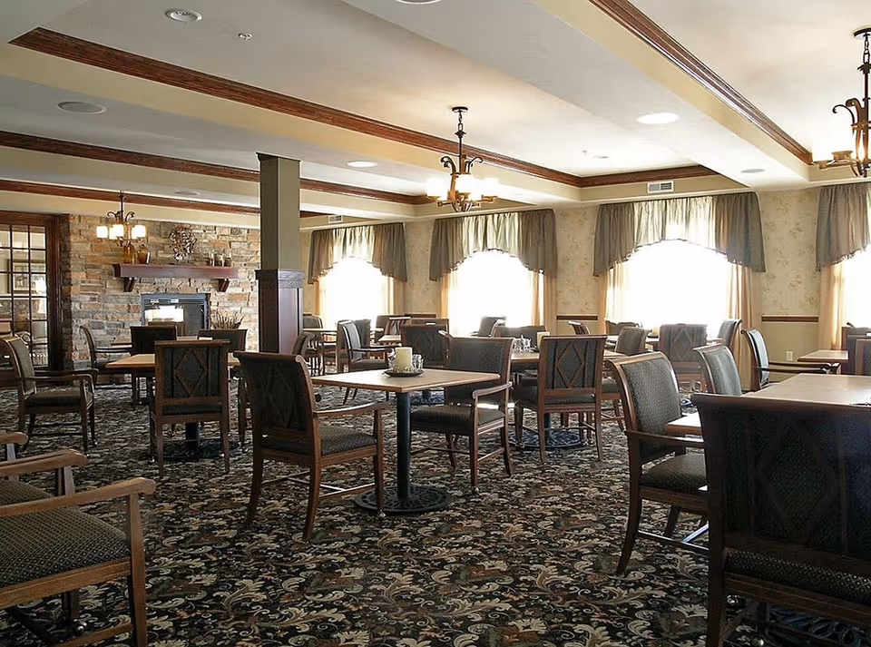 Spacious dining room with multiple wooden tables and chairs, patterned carpet, chandeliers, and large curtained windows.