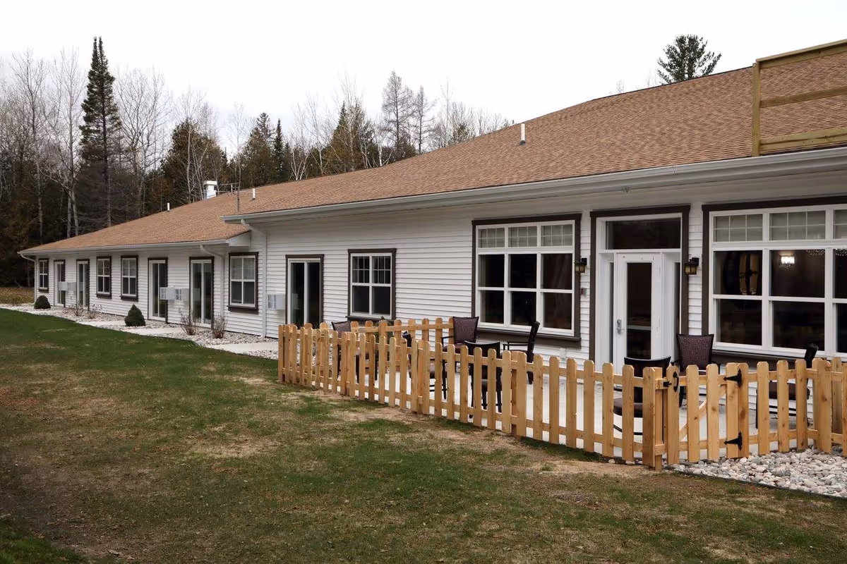 Exterior view of a single-story building with white siding and a brown roof, featuring multiple windows and doors. There is a small fenced patio area with outdoor chairs and tables. The building is surrounded by a grassy area and trees in the background.
