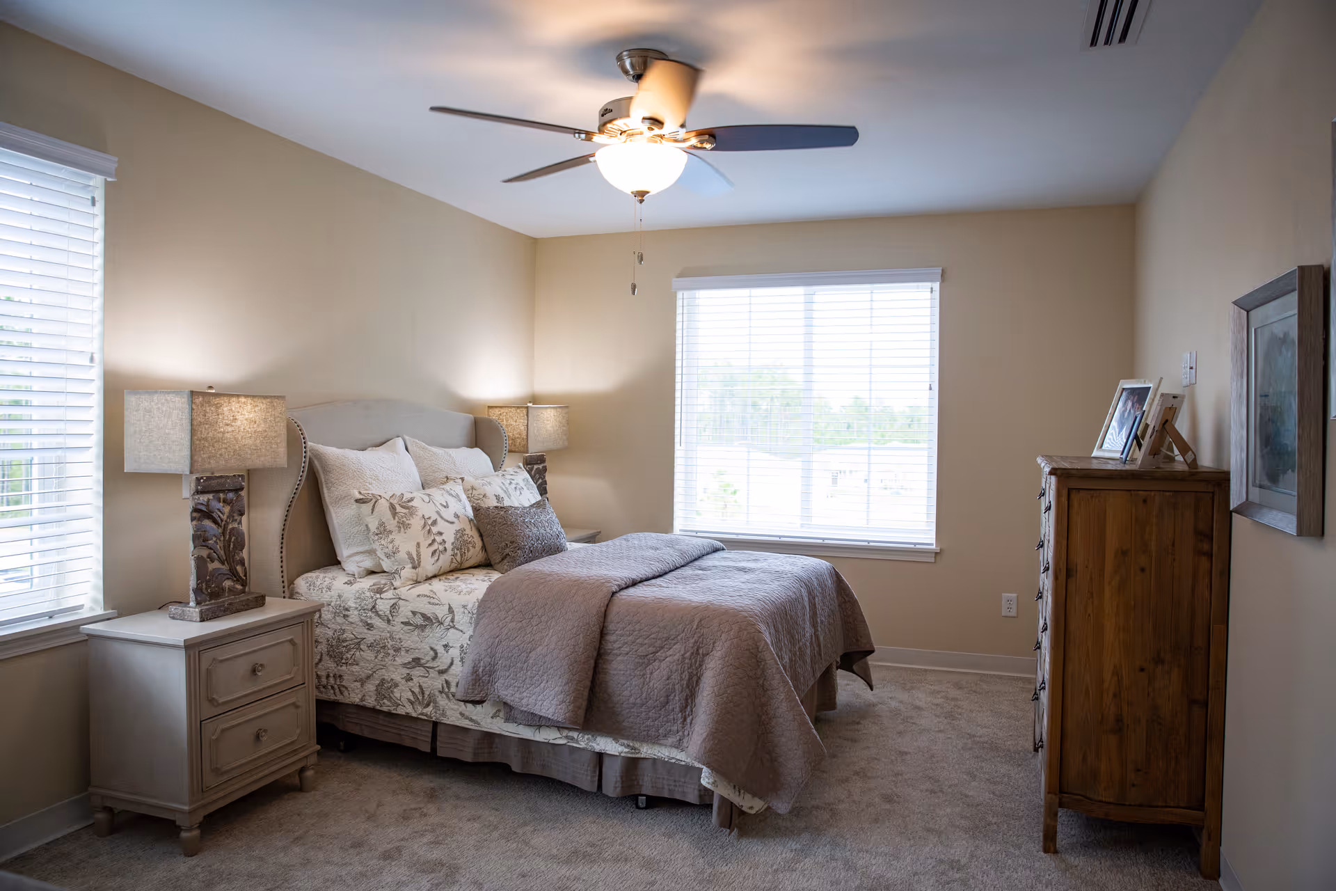 A cozy bedroom with a neatly made bed featuring floral and solid pillows, two bedside tables each with a lamp, a wooden dresser with framed photos on top, a ceiling fan with light, and two windows with white blinds letting in natural light.