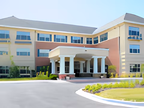 Front exterior view of a three-story senior living facility building with a covered entrance supported by white columns, surrounded by landscaped greenery and a curved driveway.