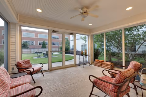 A screened-in porch area with four cushioned armchairs arranged around the perimeter. The porch has a ceiling fan and recessed lighting. Outside the screened doors, there is a grassy courtyard with a fire pit and seating. Trees and shrubs are visible around the porch and courtyard.