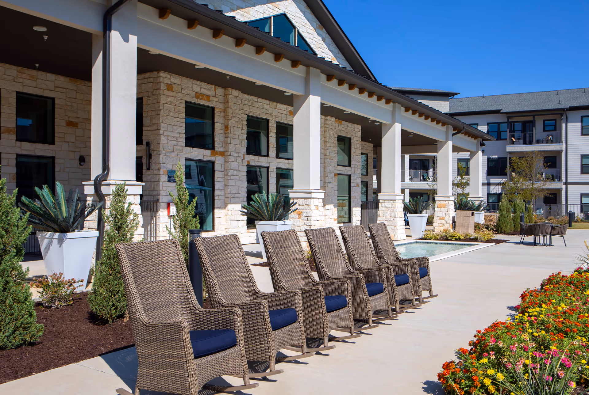 Outdoor patio area at Solea Keller featuring a row of wicker chairs with blue cushions lined up on a concrete walkway. The building has stone walls and large windows, with potted plants and landscaped flower beds nearby under a clear blue sky.