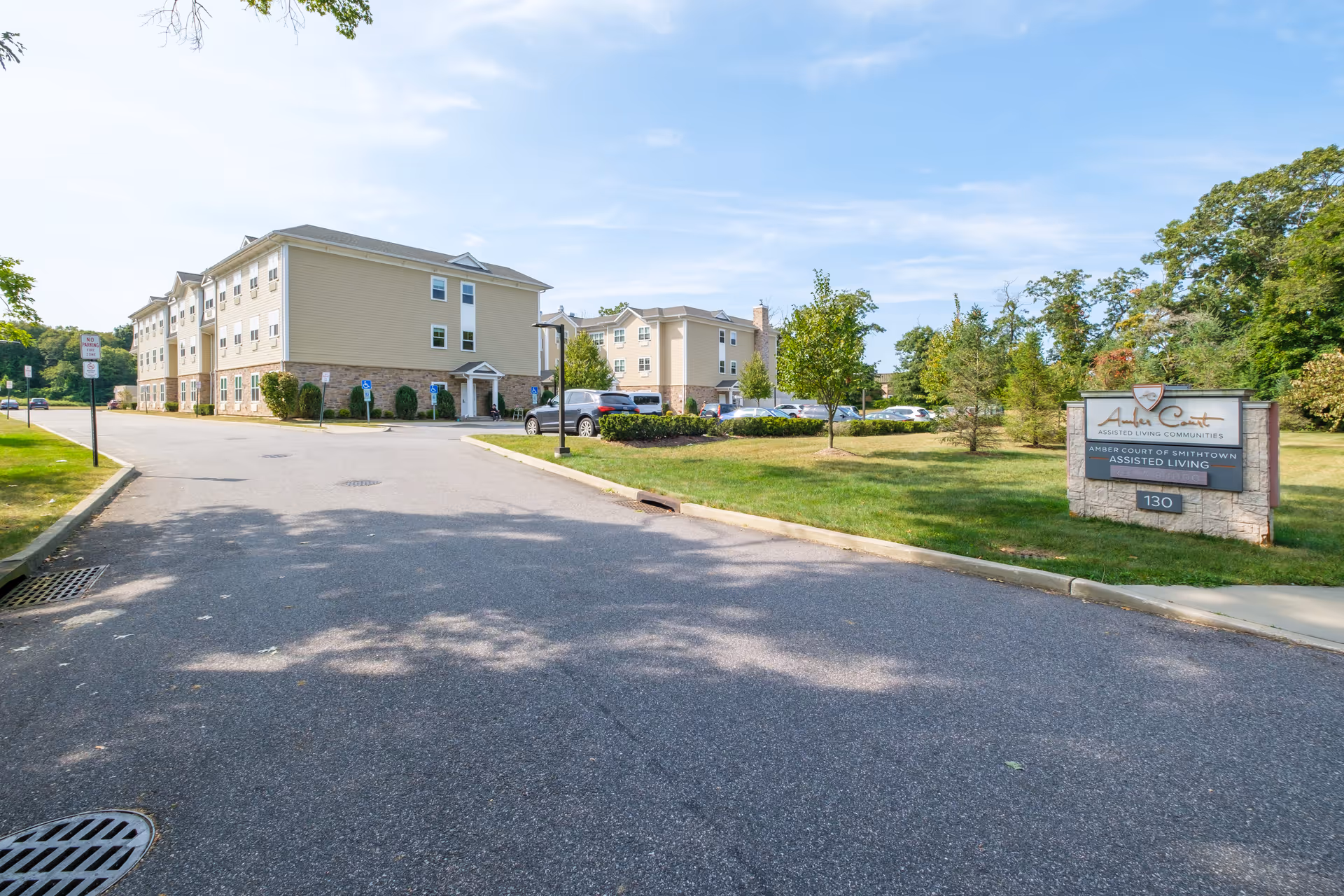 Exterior view of Amber Court assisted living community showing a three-story beige building with multiple windows, a parking lot with cars, a grassy area with trees, and a sign that reads 'Amber Court Assisted Living Communities Amber Court of Smithtown Assisted Living 130'. The sky is clear with some clouds.