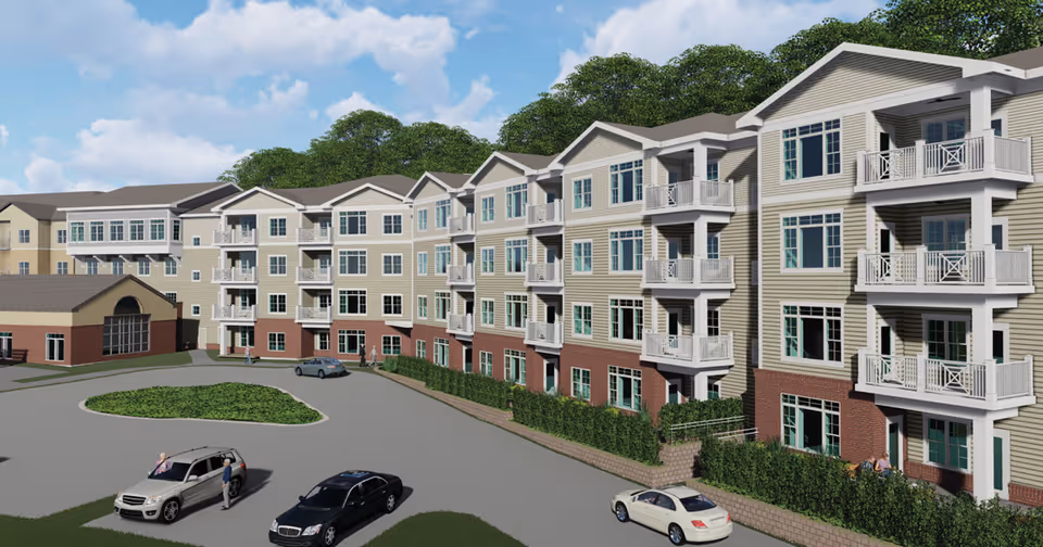Exterior view of a multi-story senior living facility named Normandie Ridge with beige siding and brick accents. The building features multiple balconies and large windows. Several cars are parked in the driveway area in front of the building, with a few people visible near the vehicles. Trees and a partly cloudy sky are in the background.