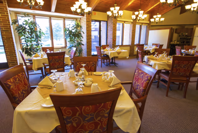 A dining room with multiple tables covered in white tablecloths, each set with folded napkins, cups, plates, and glassware. The room features wooden chairs with red and gold patterned upholstery, large windows letting in natural light, brick pillars, and chandeliers hanging from a wooden ceiling.