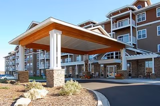 Exterior view of a multi-story senior living facility with a covered entrance supported by white columns and stone bases. The building has multiple balconies and large windows, with landscaping including rocks and shrubs near the driveway.