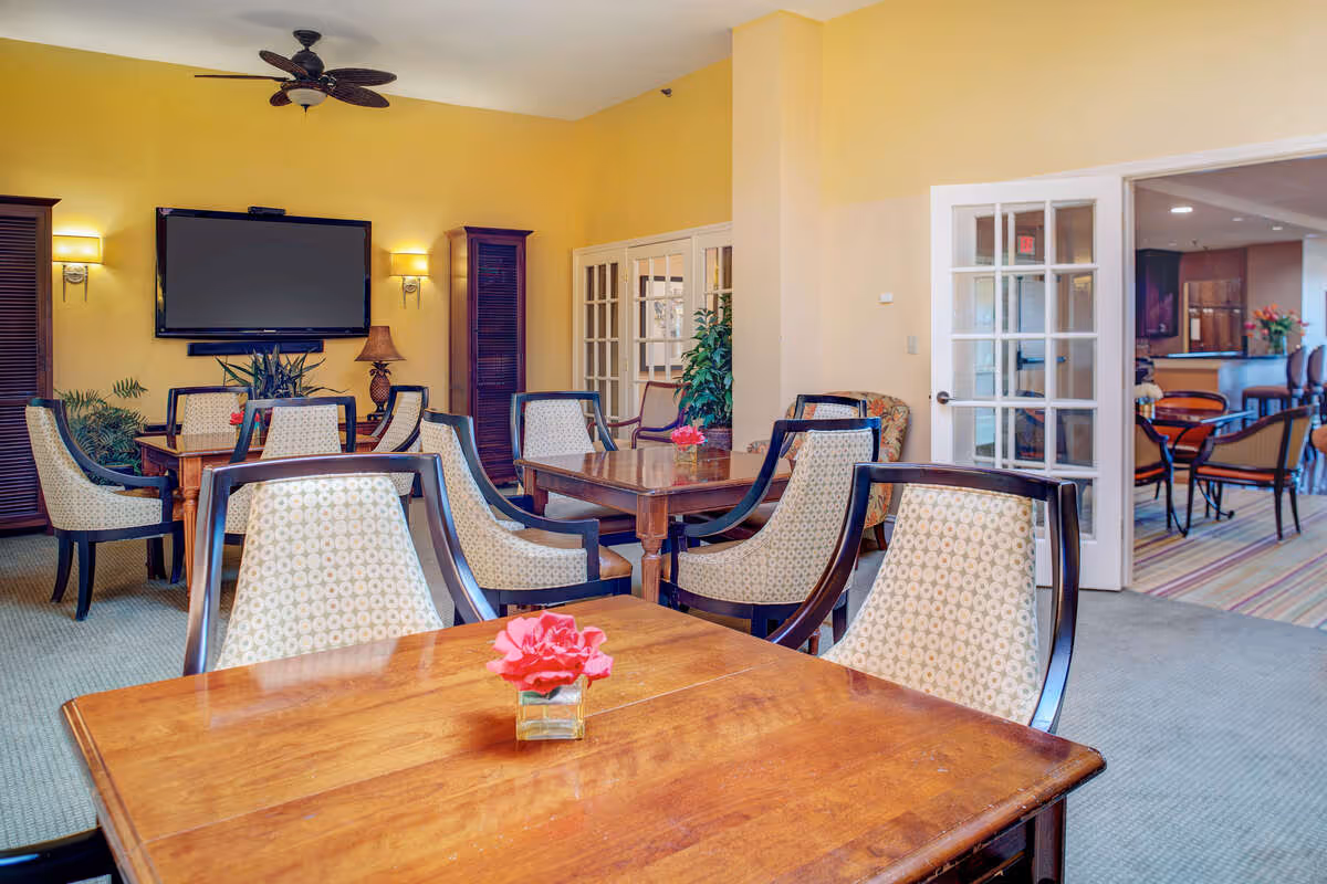 A bright and inviting dining room with wooden tables and cushioned chairs featuring patterned upholstery. A small vase with a pink flower is placed on the nearest table. The walls are painted yellow, and there is a large flat-screen TV mounted on the wall flanked by two wall sconces. French doors lead to another room with more seating and a bar area visible in the background.