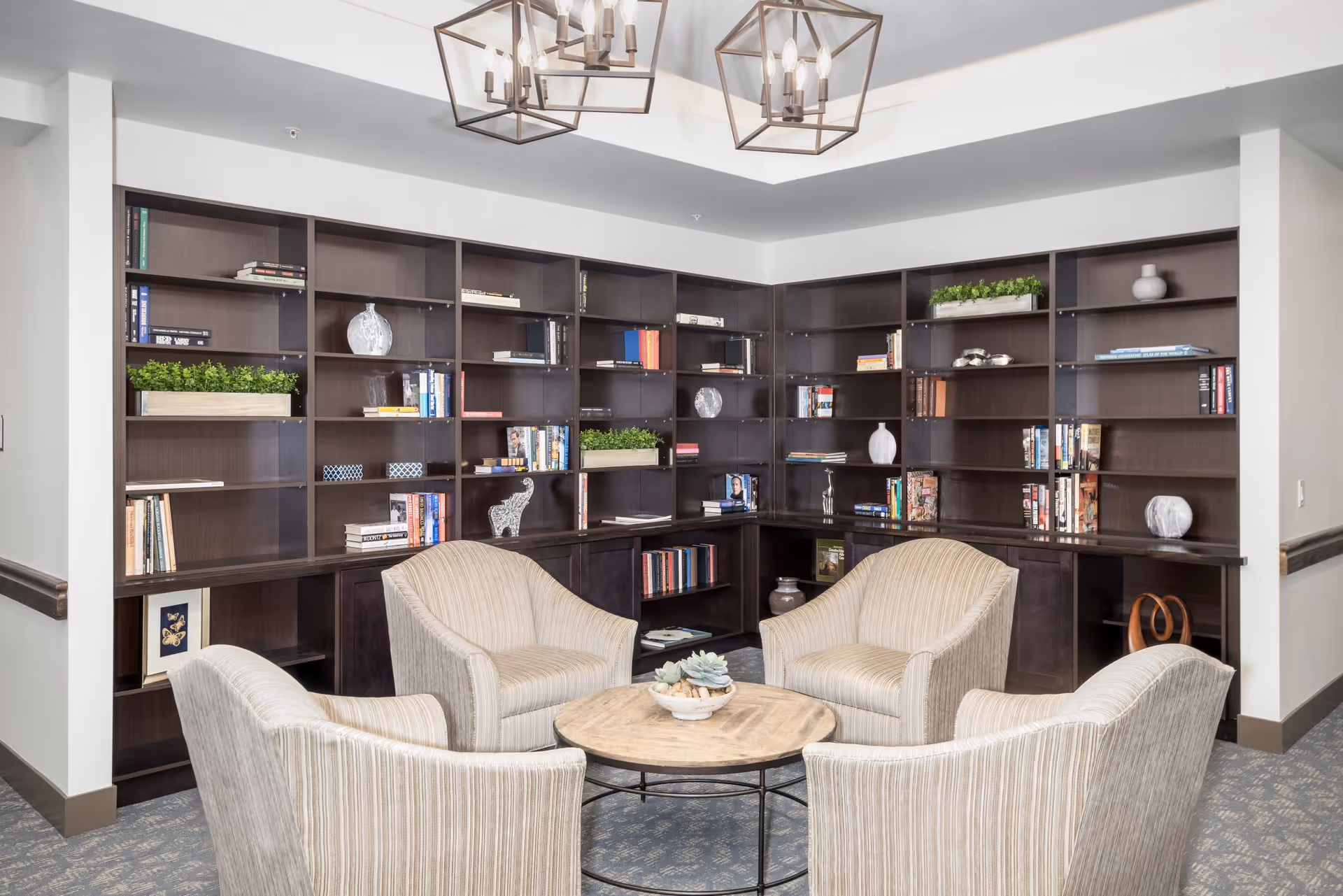 A cozy seating area in a senior living facility with four beige striped armchairs arranged around a round wooden coffee table. Behind the seating area is a large dark wood bookshelf filled with books, decorative items, and small plants. The ceiling features two modern geometric light fixtures.