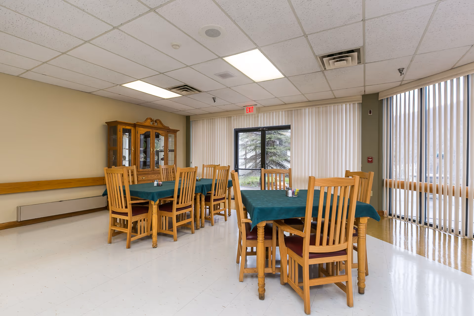 A dining room with two wooden tables covered with green tablecloths, each surrounded by wooden chairs with red cushions. A wooden china cabinet is against the beige wall. Large windows with vertical blinds allow natural light to enter the room.