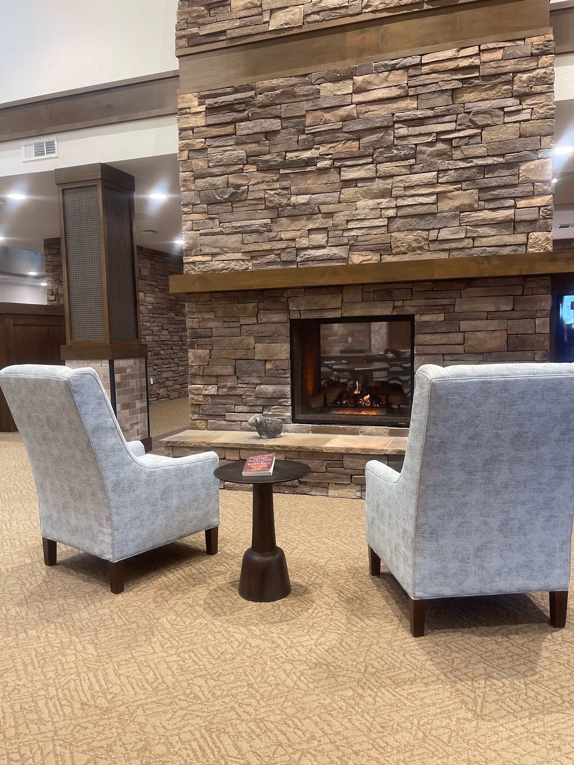 Two light gray upholstered armchairs facing a stone fireplace with a small round dark wooden table between them. A book and a decorative item are on the table. The room has beige patterned carpet and wood and stone accents on the walls.