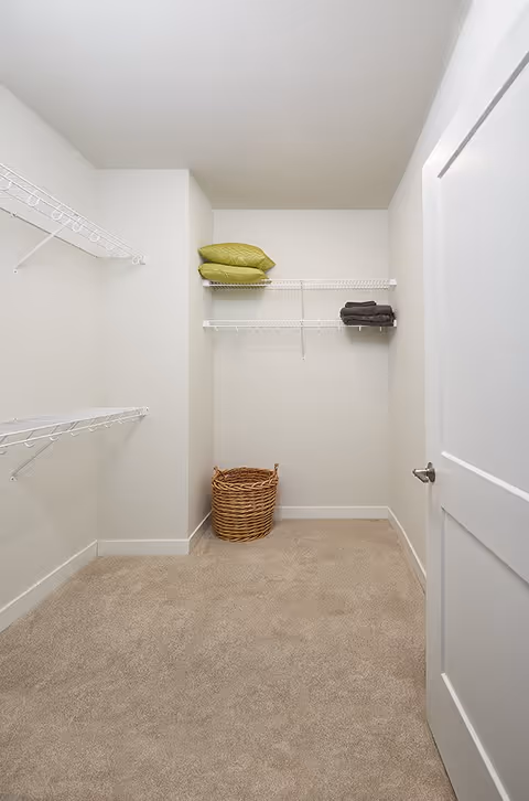 A small walk-in closet with beige carpet, white walls, and white wire shelving. There are two green pillows and two folded dark towels on the shelves, and a woven basket on the floor. The closet door is partially open.