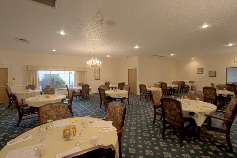 A spacious dining room with multiple round tables covered with cream-colored tablecloths. Each table is set with glassware, napkins, menus, and small decorative pumpkins. The chairs have floral upholstery. The room has a patterned carpet, cream-colored walls, a large window with curtains, and a chandelier hanging from the ceiling.