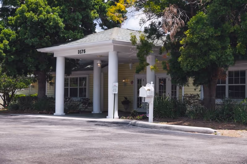 Exterior view of a senior living facility entrance with white columns supporting a small roof overhang. The building is light yellow with white trim, surrounded by green trees and bushes. A white mailbox is visible near the driveway, and the number 3175 is displayed above the entrance.