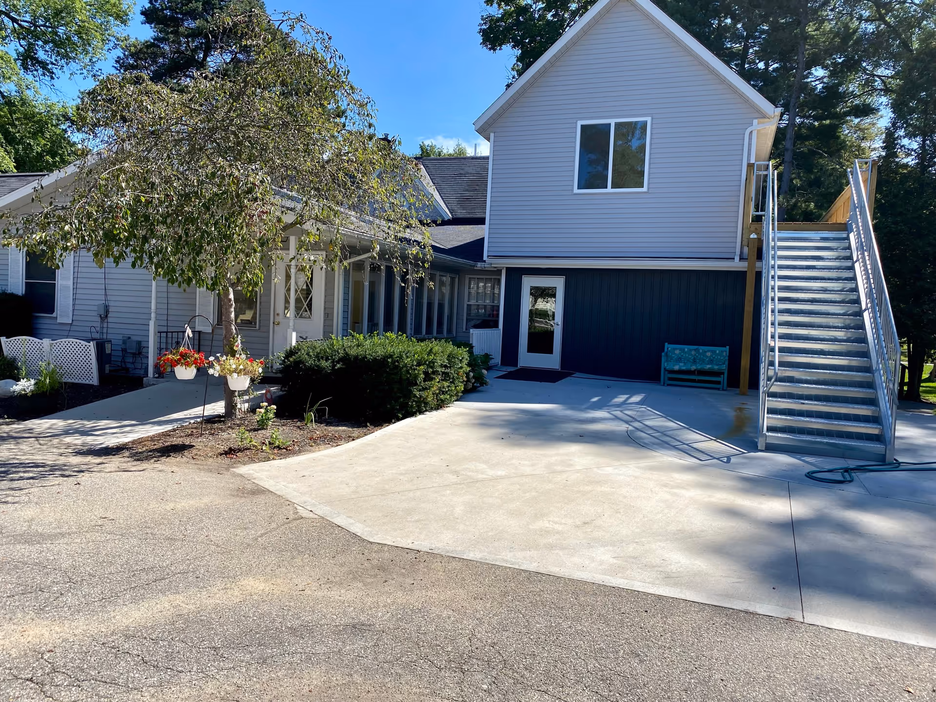 Exterior view of a senior living facility building with gray siding and a white door. There is a concrete driveway and a metal staircase leading to the upper floor. A tree with hanging flower pots and some bushes are visible near the entrance. The sky is clear and blue.