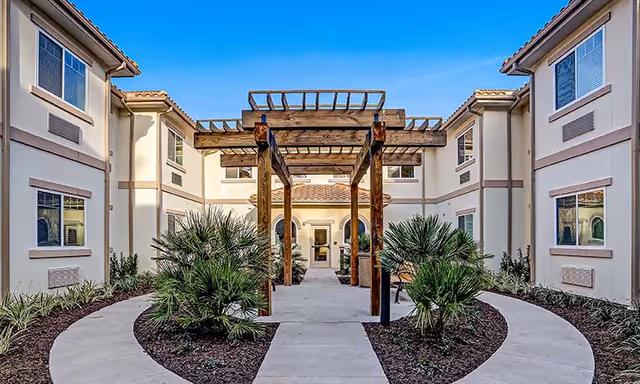 Outdoor courtyard area at HarborChase of Daytona Beach featuring a wooden pergola structure in the center, surrounded by landscaped plants and a curved concrete walkway leading to the entrance of a two-story building with multiple windows.