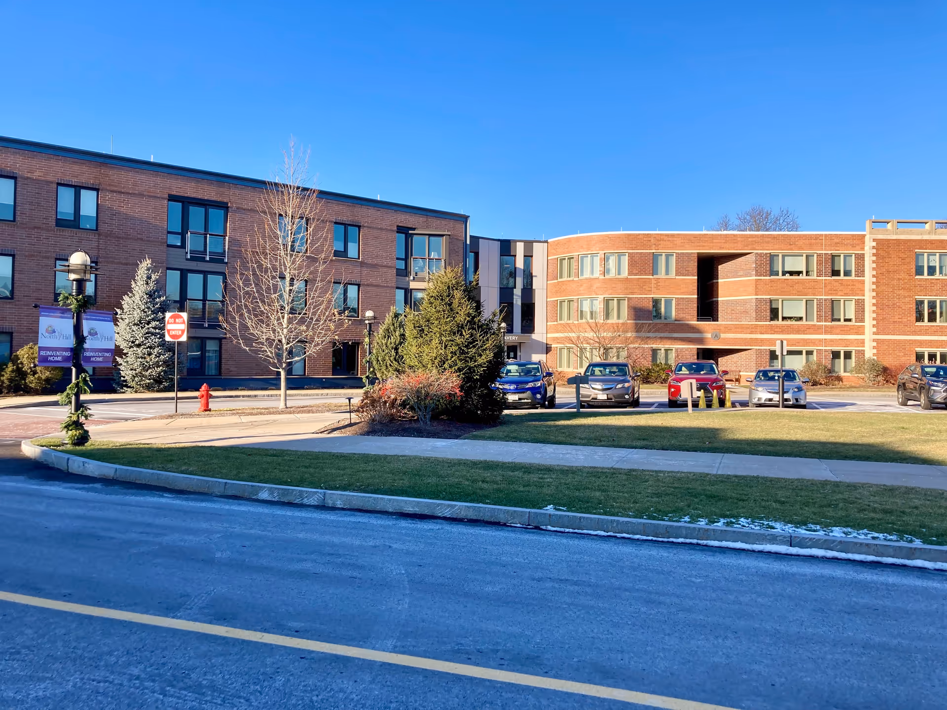 Front exterior of a brick multi-story retirement community building with parked cars, lawn, and clear blue sky.