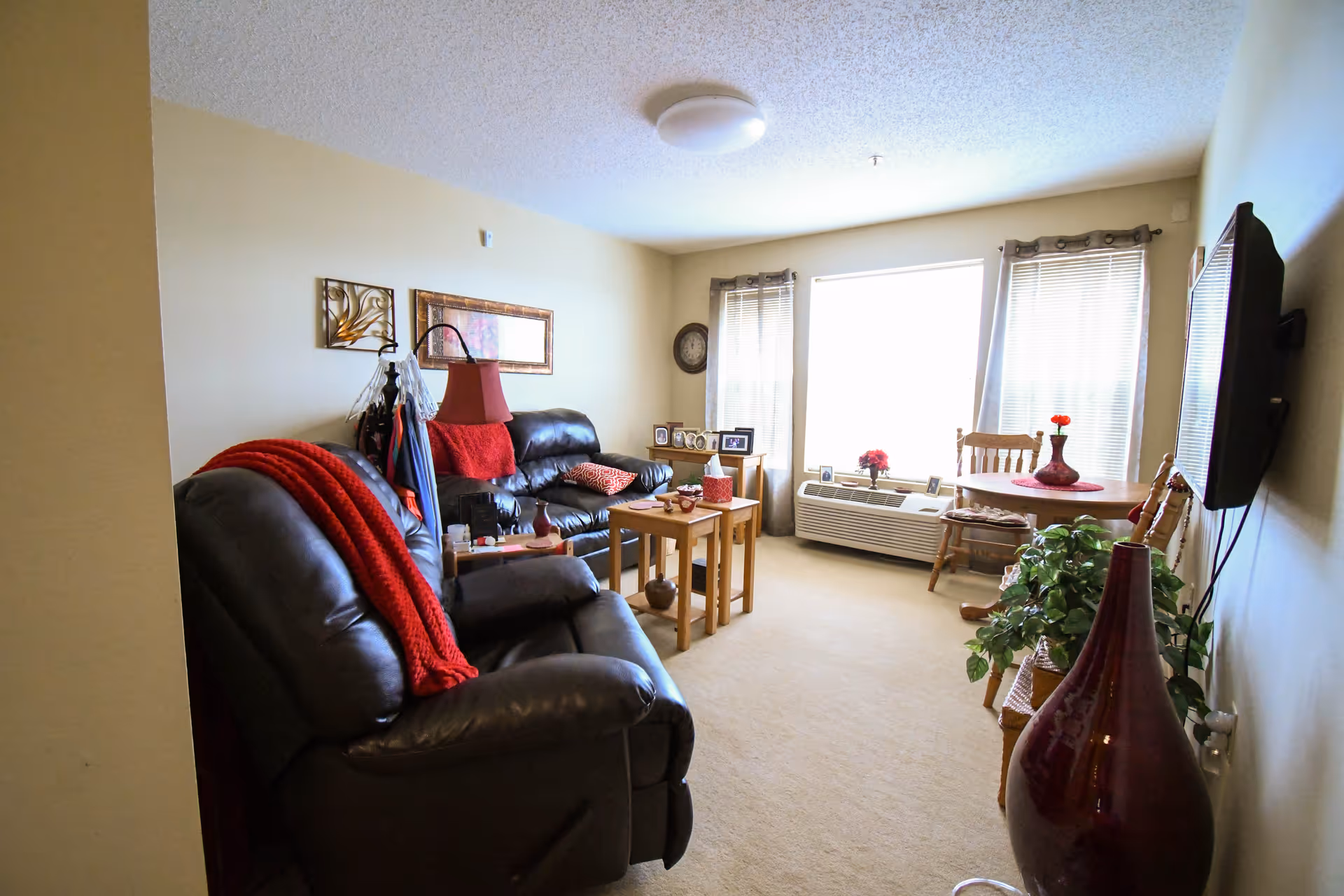 A cozy living room with beige walls and carpet, featuring two dark leather sofas with red blankets and pillows, a small wooden table with decorative items, a wall-mounted flat-screen TV, a round wooden dining table with chairs near a large window with blinds and curtains, and various framed photos and plants around the room.