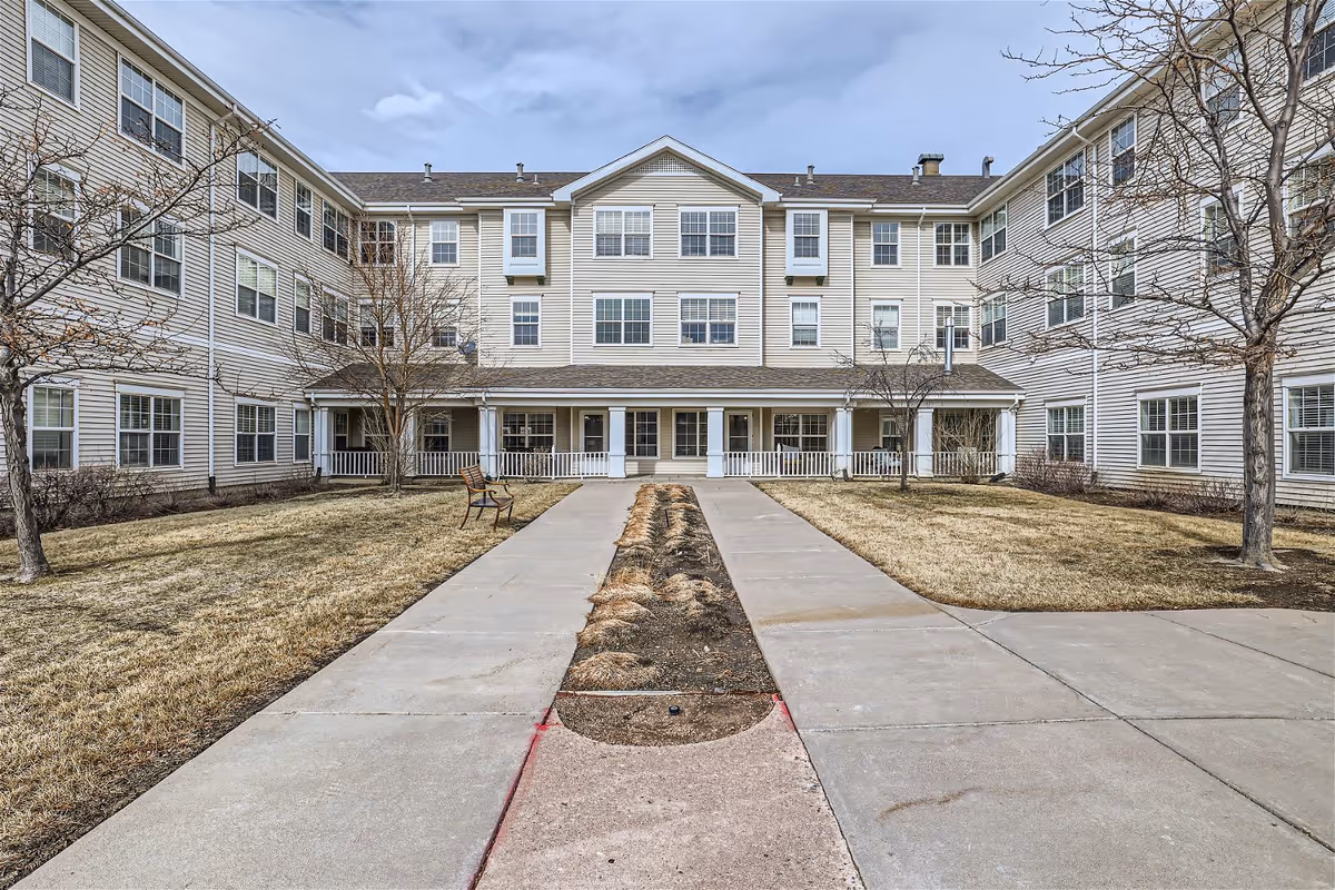 Exterior view of a three-story senior living facility building with beige siding and multiple windows. There is a paved walkway leading to the entrance, flanked by dry grass and leafless trees. A bench is placed along the walkway near the entrance.