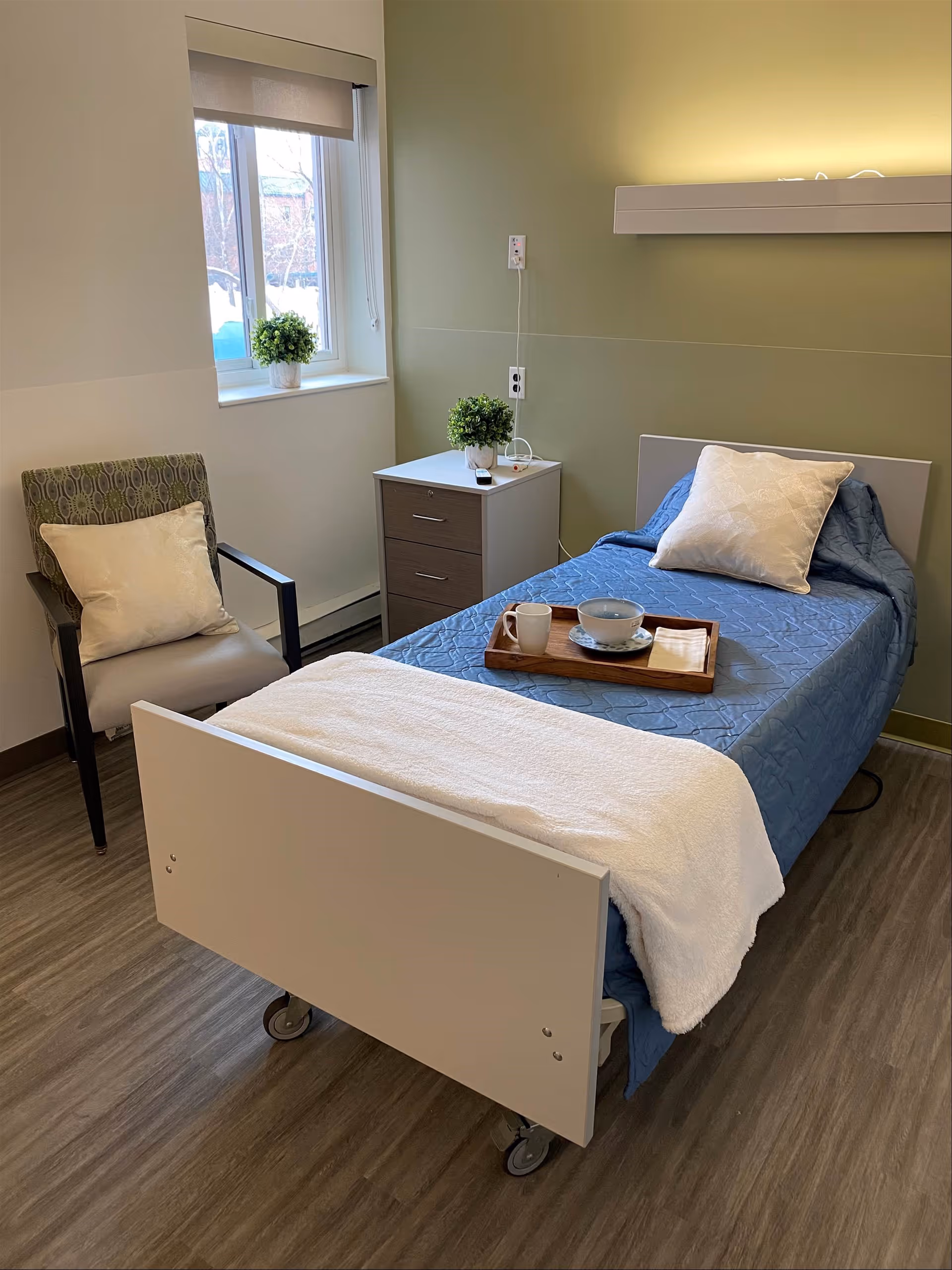A small resident bedroom with a single hospital-style bed made with blue bedding and a tray, a nightstand, armchair, and a window with potted plants.