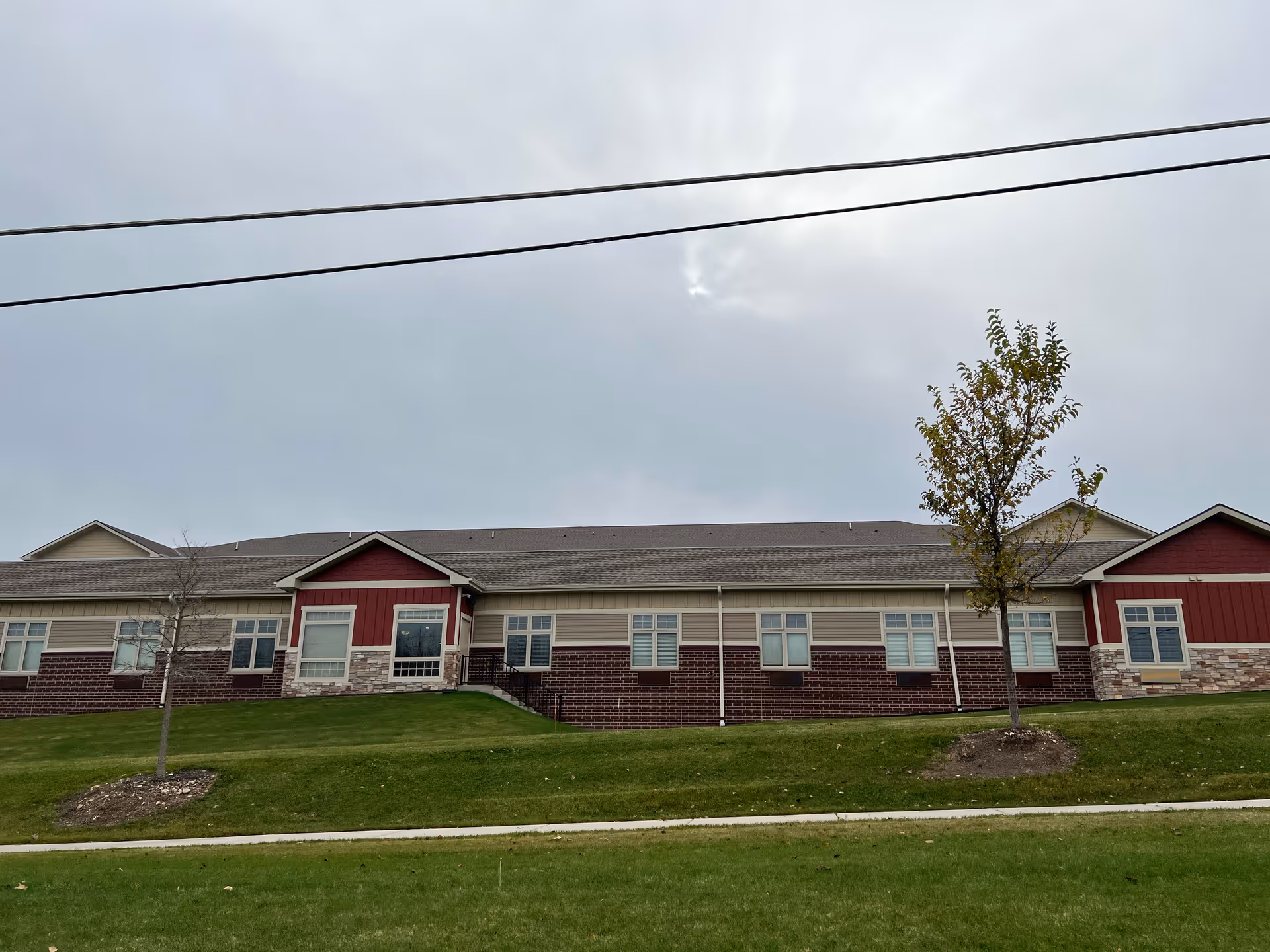 Single-story senior living building with red and beige siding and a brick lower facade, windows, small trees and a grassy lawn under a cloudy sky.