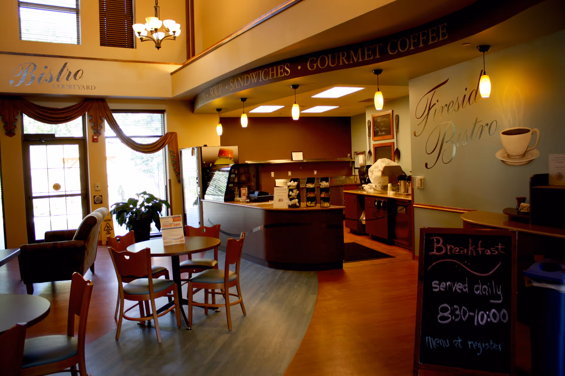 Interior view of a cozy bistro area with wooden tables and chairs, a counter for ordering food and drinks, warm lighting, and a sign on the wall that reads 'Fireside Bistro'. A chalkboard sign indicates breakfast hours from 8:30 to 10:00. Large windows and a door provide natural light, and there is a comfortable armchair near the entrance.