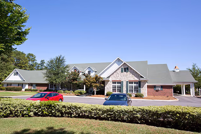 Exterior view of a single-story senior living facility building with a combination of brick and stone facade, multiple windows, and a green roof under a clear blue sky. There are two cars parked in front of the building and neatly trimmed bushes and trees surrounding the area.