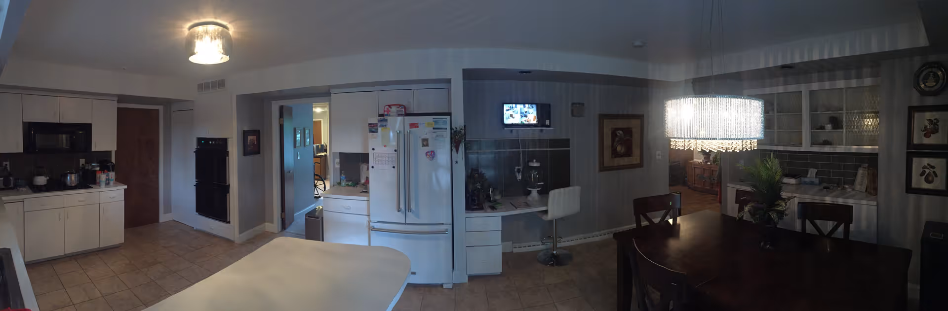 A spacious kitchen and dining area in a senior living facility. The kitchen features white cabinets, a refrigerator, a microwave, and an oven. There is a white countertop island in the center. The dining area has a dark wooden table with chairs and a modern chandelier hanging above it. The walls are decorated with framed artwork and built-in shelves. The floor is tiled, and the lighting is warm and inviting.