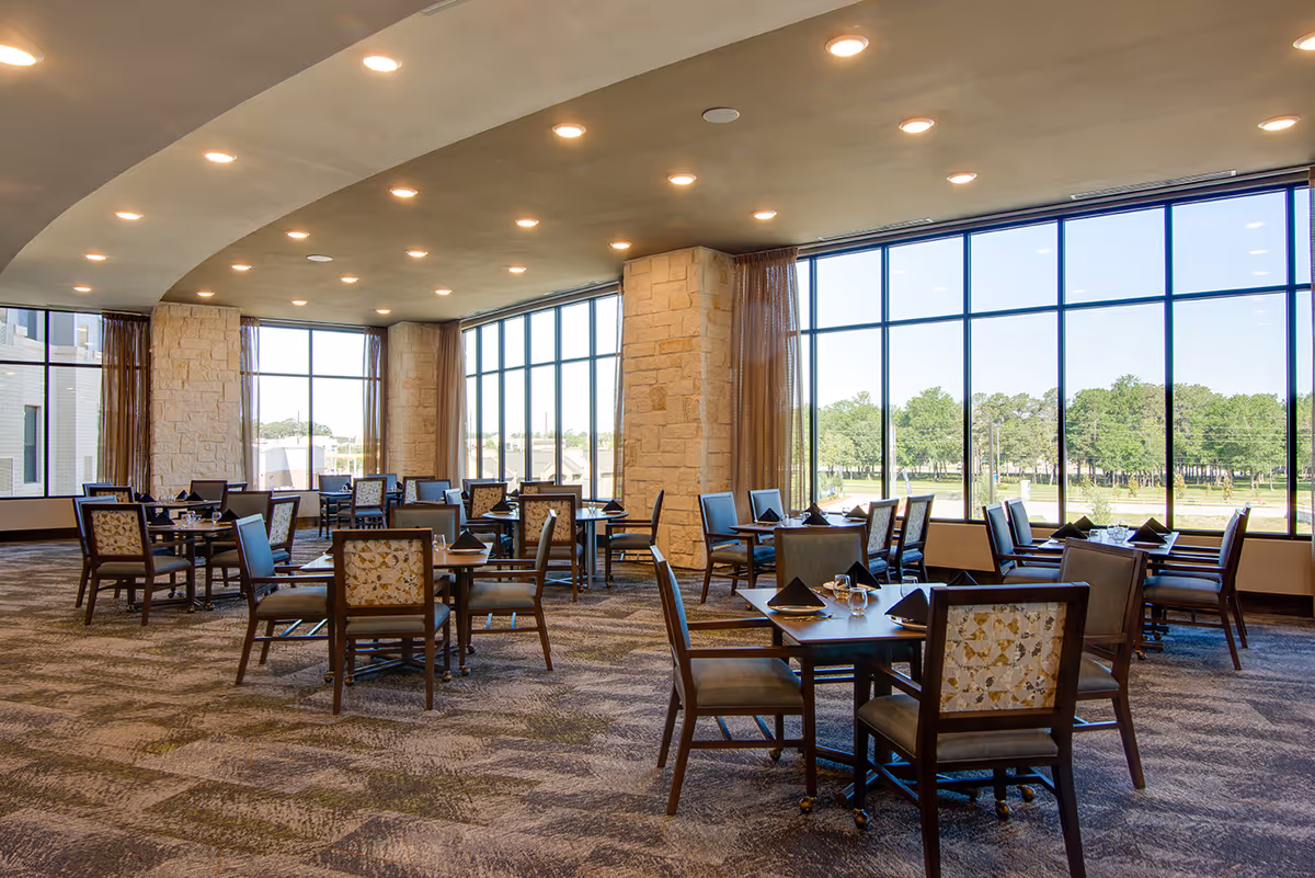A spacious dining room with multiple tables and chairs arranged neatly. The room features large floor-to-ceiling windows that provide a clear view of green trees and a bright blue sky outside. The ceiling has recessed lighting, and the walls include stone pillars and curtains.