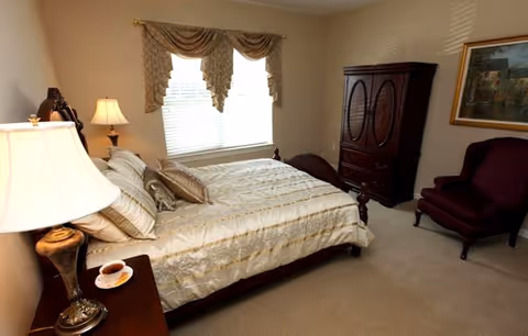 A bedroom with a large bed covered in a beige and gold patterned bedspread, two pillows, and a decorative pillow. There is a wooden nightstand with a lamp and a cup of tea on a saucer. The room has a window with beige curtains, a wooden armoire, a burgundy upholstered armchair, and a framed painting on the wall.
